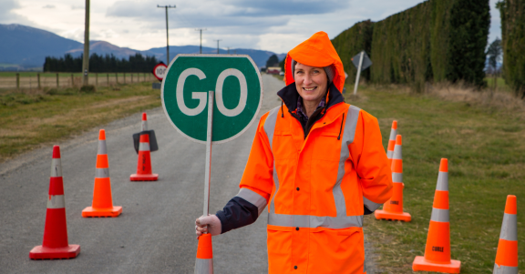 Road worker eearing a bright orange high-vis jacket hols a green sign with "GO" written in white. In the background is a road under construction with orange traffic cones dotted around.